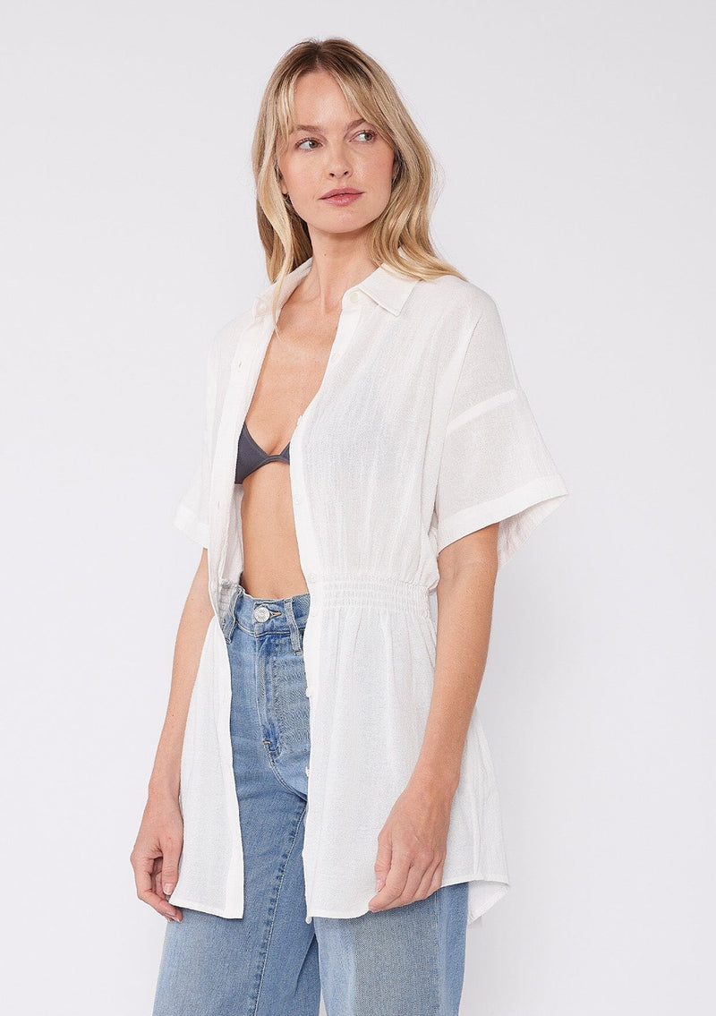 [Color: White] A half body side facing image of a blonde model wearing a white tunic shirt with a collared neckline, a button front, short sleeves with a dropped shoulder, and a smocked elastic waist. 