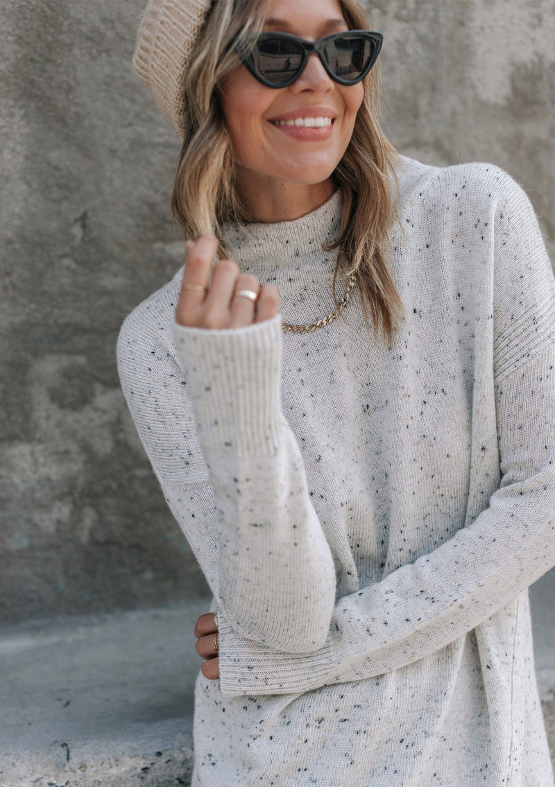 [Color: Silver] A woman standing outside wearing a mélange knit sweater dress. Featuring a mock neckline, long sleeves with contrast ribbed details, and an asymmetric front slit detail.