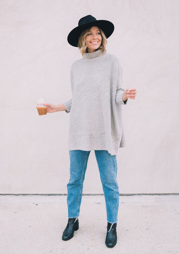 [Color: Heather Grey] A woman wearing a cable knit pullover. Featuring contrast ribbed sleeves and turtleneck, a relaxed, oversize fit and tunic style length, and flattering three quarter length dolman sleeves.