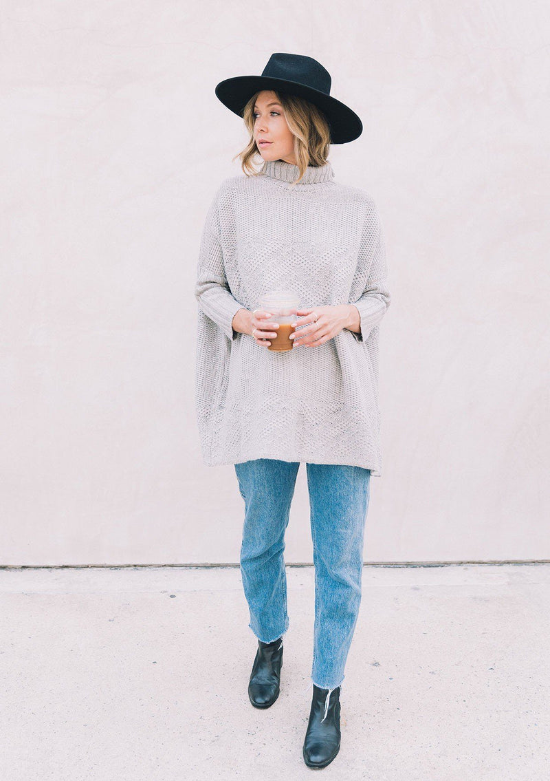 [Color: Heather Grey] A woman wearing a cable knit pullover. Featuring contrast ribbed sleeves and turtleneck, a relaxed, oversize fit and tunic style length, and flattering three quarter length dolman sleeves.