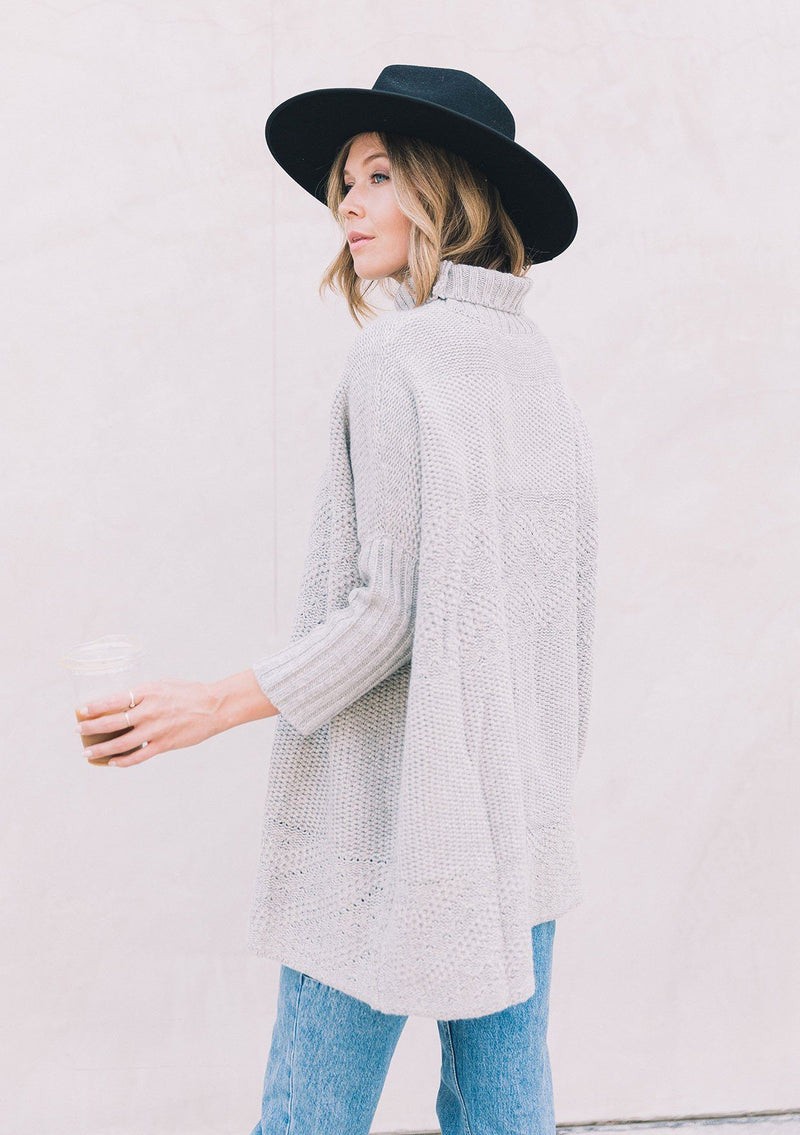 [Color: Heather Grey] A woman wearing a cable knit pullover. Featuring contrast ribbed sleeves and turtleneck, a relaxed, oversize fit and tunic style length, and flattering three quarter length dolman sleeves.