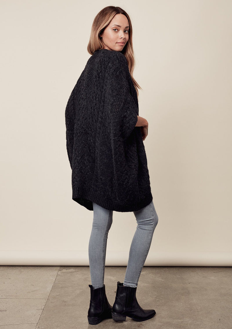 [Color: Charcoal] A side image of a brunette model wearing a cocoon style poncho in dark grey black. This cable knit poncho features cozy short sleeves, a v-neckline, zipper front, and a rib trimmed hem. A layering essential for the fall season. 