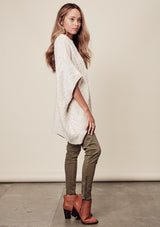 [Color: Heather Petal] A side image of a brunette model wearing a cocoon style poncho in light beige. This cable knit poncho features cozy short sleeves, a v-neckline, zipper front, and a rib trimmed hem. A layering essential for the fall season. 