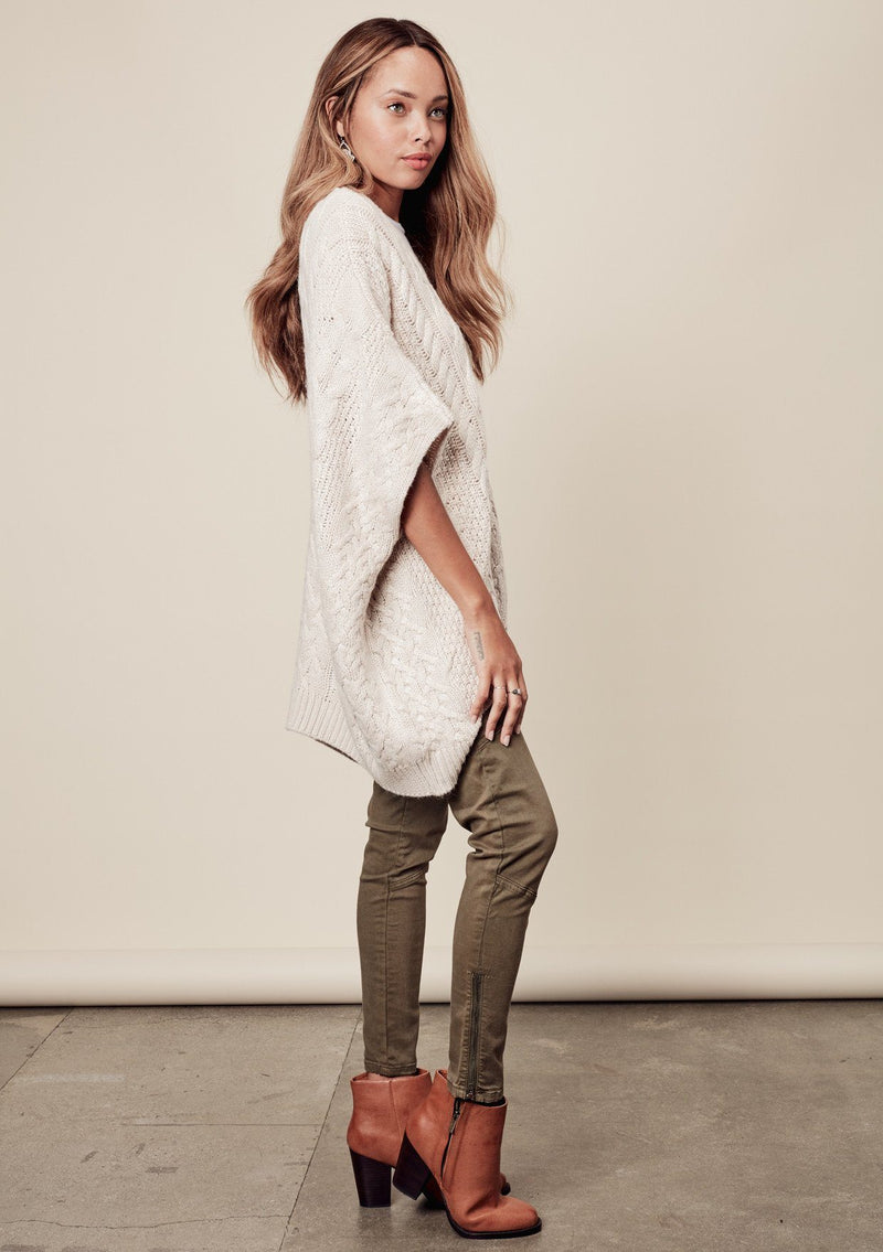 [Color: Heather Petal] A side image of a brunette model wearing a cocoon style poncho in light beige. This cable knit poncho features cozy short sleeves, a v-neckline, zipper front, and a rib trimmed hem. A layering essential for the fall season. 