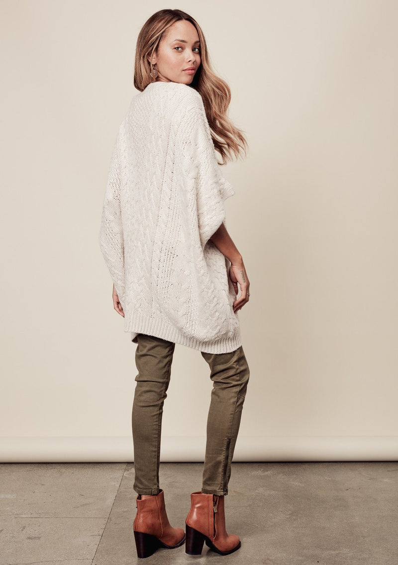 [Color: Heather Petal] A side image of a brunette model wearing a cocoon style poncho in light beige. This cable knit poncho features cozy short sleeves, a v-neckline, zipper front, and a rib trimmed hem. A layering essential for the fall season. 