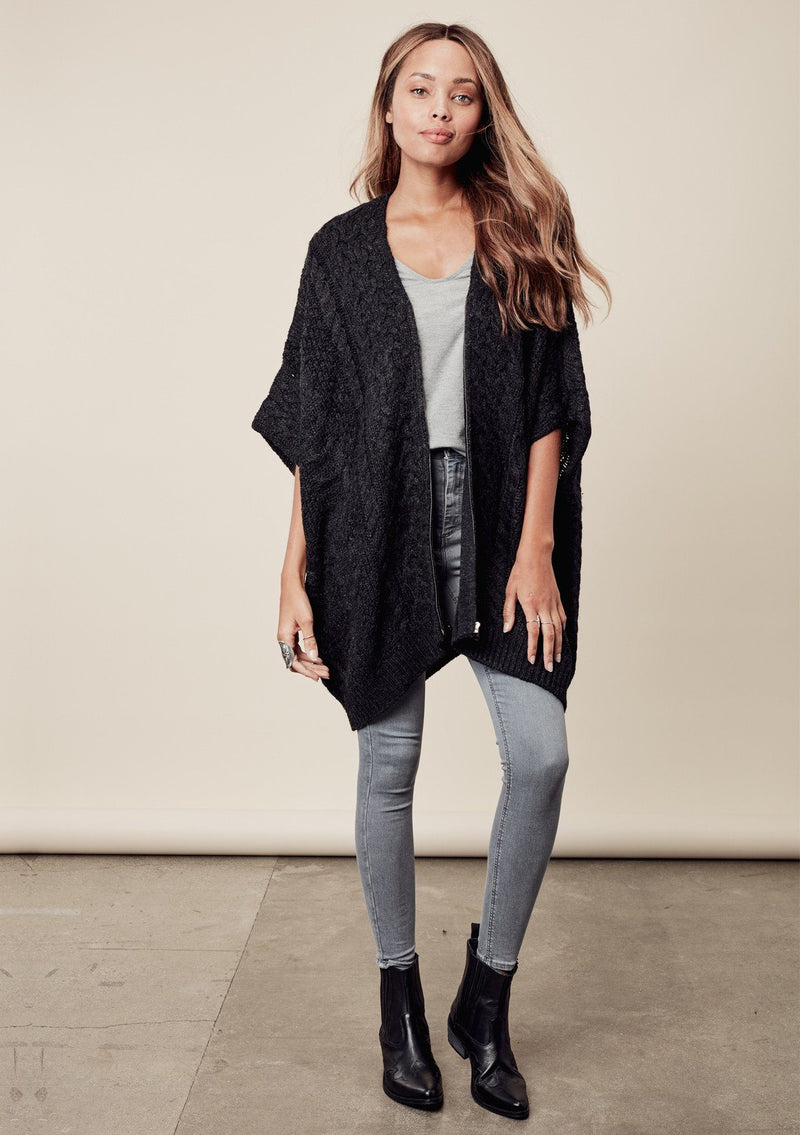 [Color: Charcoal] A front image of a brunette model wearing a cocoon style poncho in dark grey black. This cable knit poncho features cozy short sleeves, a v-neckline, zipper front, and a rib trimmed hem. A layering essential for the fall season. 