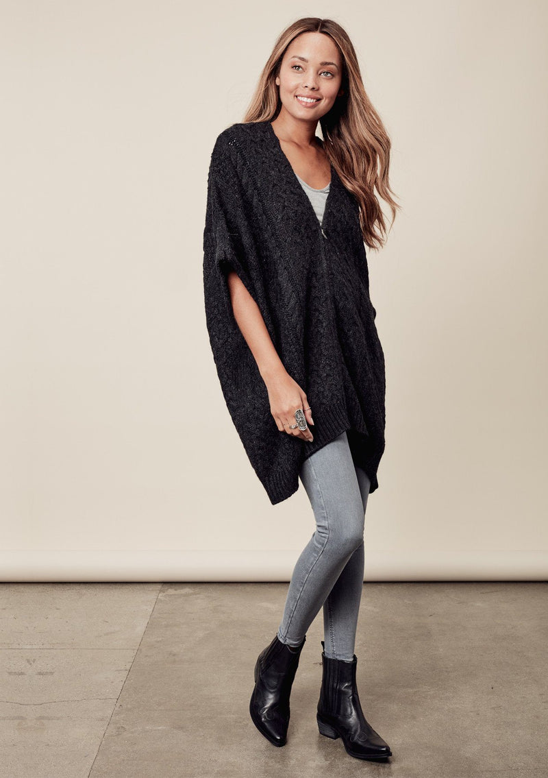 [Color: Charcoal] A front image of a brunette model wearing a cocoon style poncho in dark grey black. This cable knit poncho features cozy short sleeves, a v-neckline, zipper front, and a rib trimmed hem. A layering essential for the fall season. 