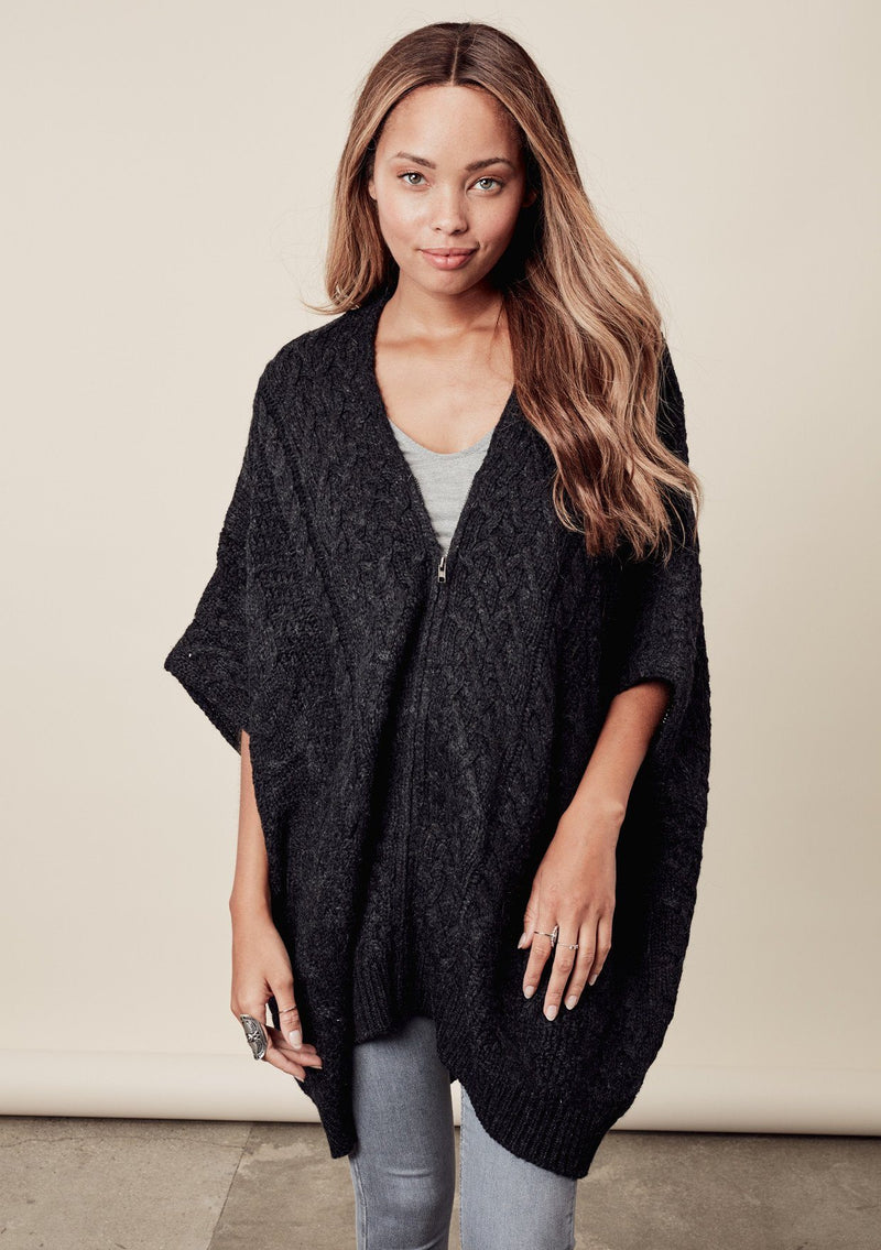 [Color: Charcoal] A front image of a brunette model wearing a cocoon style poncho in dark grey black. This cable knit poncho features cozy short sleeves, a v-neckline, zipper front, and a rib trimmed hem. A layering essential for the fall season. 