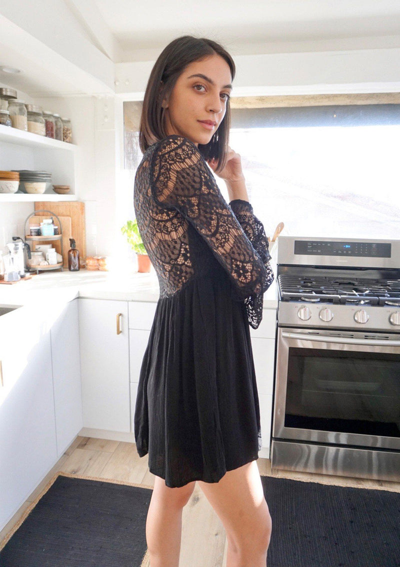 [Color: Black] A brunette woman wearing a sheer lace mini dress. Featuring a feminine v neckline, a button front detail, an empire waist, long sheer lace bell sleeves, and a sexy sheer lace back detail.