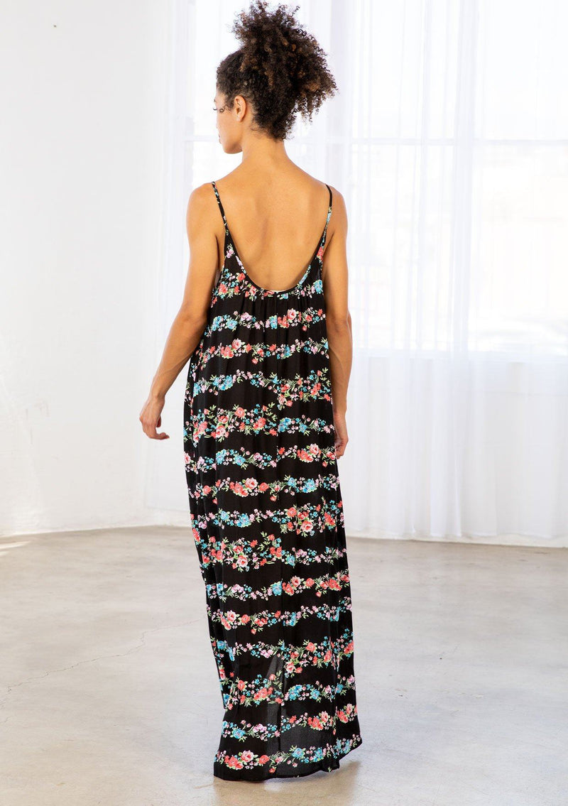 [Color: Black Combo] A brunette model wearing a sleeveless scoop neck maxi dress in black floral print. A colorful flowy floral dress with a high low hemline and relaxed fit. 