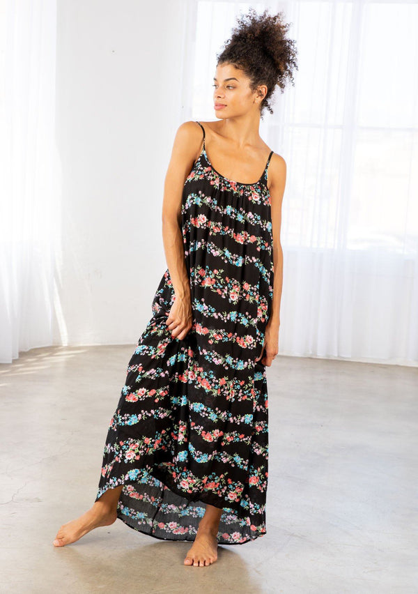 [Color: Black Combo] A brunette model wearing a sleeveless scoop neck maxi dress in black floral print. A colorful flowy floral dress with a high low hemline and relaxed fit. 