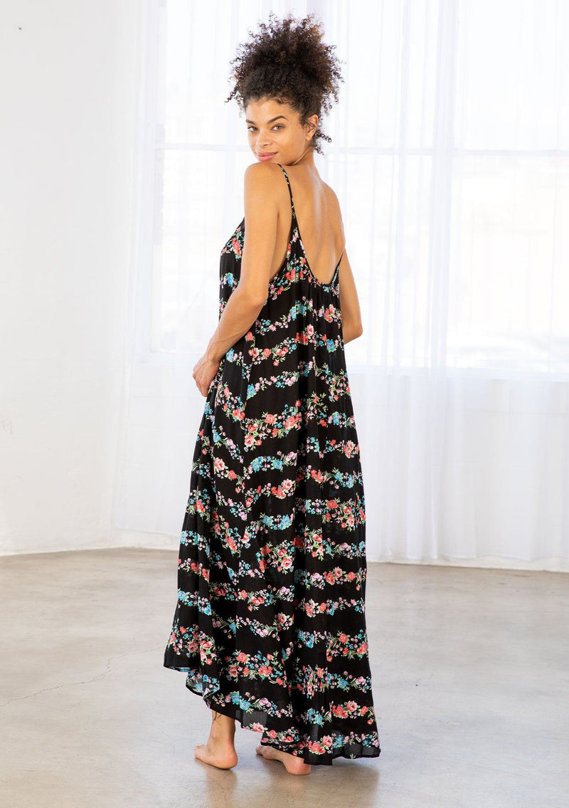 [Color: Black Combo] A brunette model wearing a sleeveless scoop neck maxi dress in black floral print. A colorful flowy floral dress with a high low hemline and relaxed fit. 