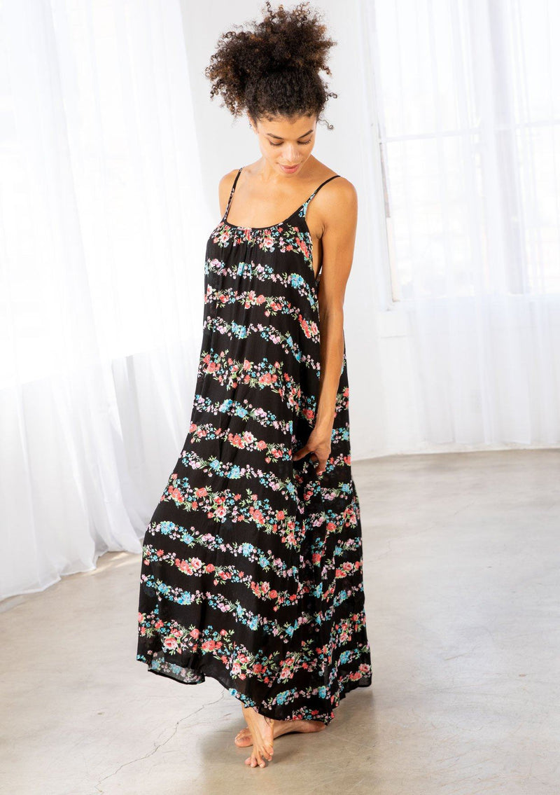 [Color: Black Combo] A brunette model wearing a sleeveless scoop neck maxi dress in black floral print. A colorful flowy floral dress with a high low hemline and relaxed fit. 