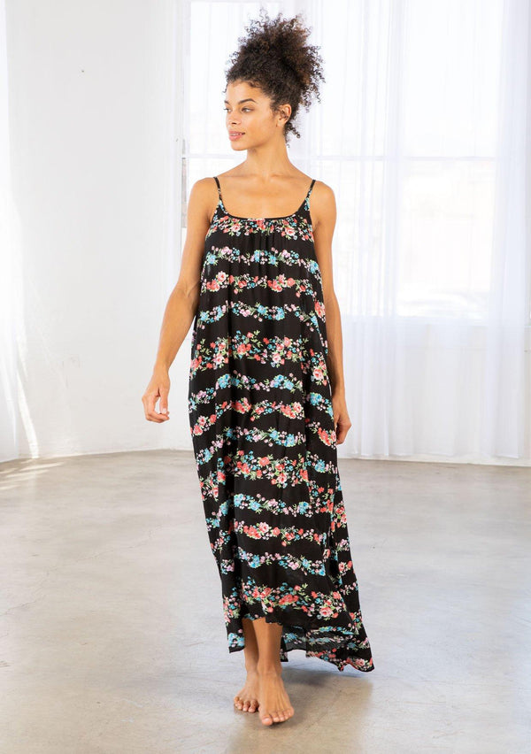 [Color: Black Combo] A brunette model wearing a sleeveless scoop neck maxi dress in black floral print. A colorful flowy floral dress with a high low hemline and relaxed fit. 