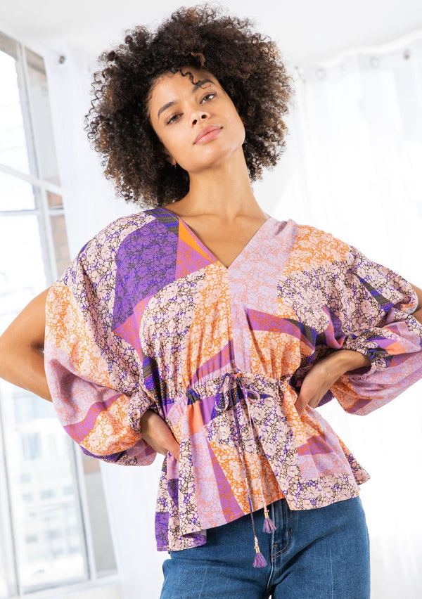 [Color: Pink/Coral] A model wearing a pink and coral floral patchwork print blouse. With long voluminous split sleeves, an elastic wrist cuff, a drawstring waist, and an open back with tassel tie closure. 