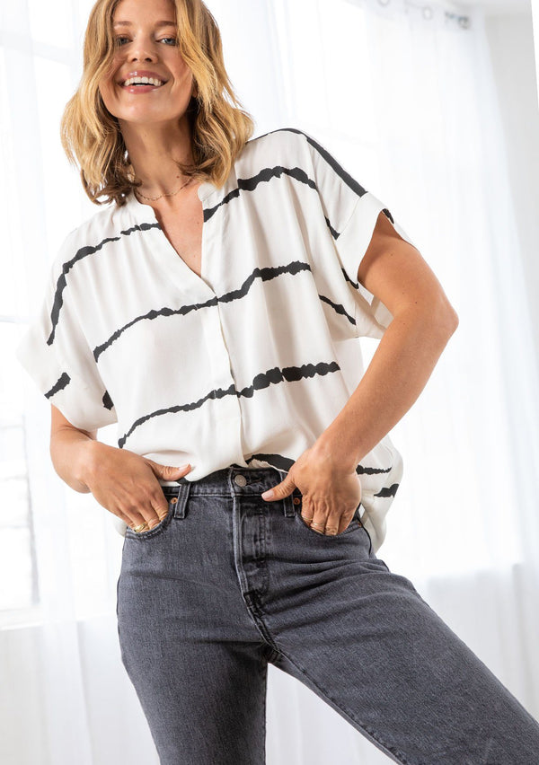 [Color: Off White/Black] A model wearing an off white popover top with abstract black stripes. Featuring a short sleeve and banded collar. 