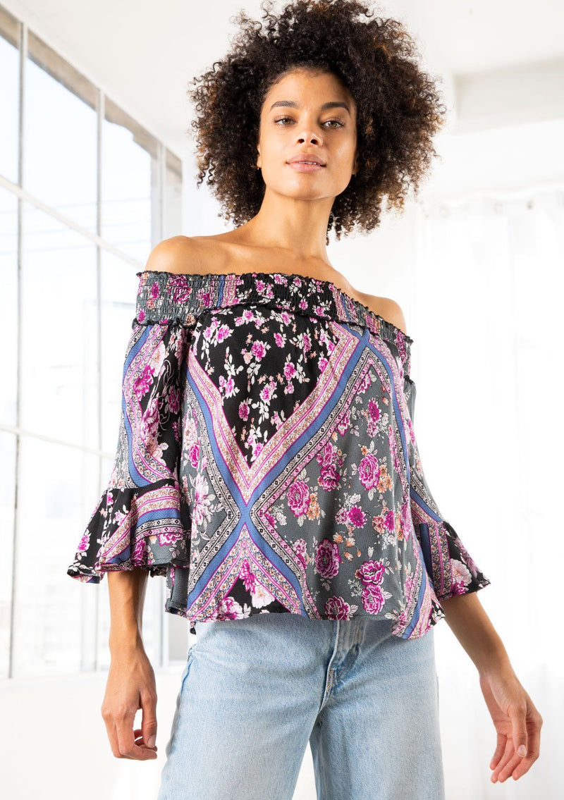 [Color: Black/Orchid] A model wearing a floral patchwork blouse. With three quarter length sleeves and a flounced cuff, a billowy relaxed fit, and a smocked elastic off shoulder detail. 