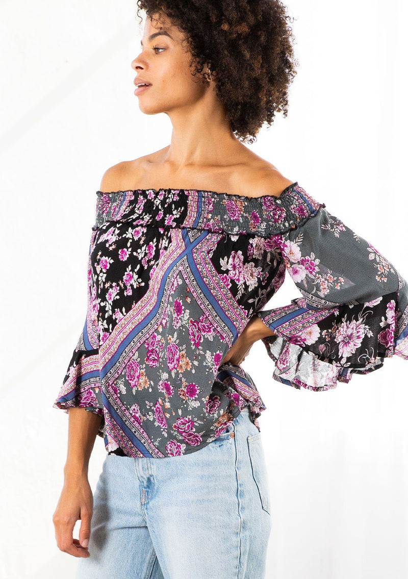 [Color: Black/Orchid] A model wearing a floral patchwork blouse. With three quarter length sleeves and a flounced cuff, a billowy relaxed fit, and a smocked elastic off shoulder detail. 