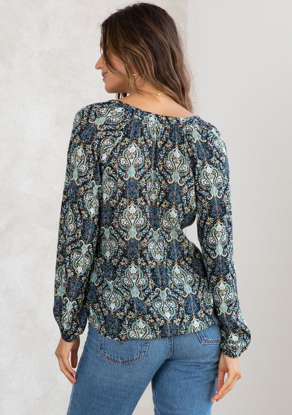 [Color: Black/Teal] A model wearing a black and teal paisley print peasant blouse with gold lurex thread details. With long voluminous sleeves, a self covered button front, and a button loop trimmed v neckline. 