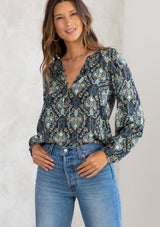 [Color: Black/Teal] A model wearing a black and teal paisley print peasant blouse with gold lurex thread details. With long voluminous sleeves, a self covered button front, and a button loop trimmed v neckline. 