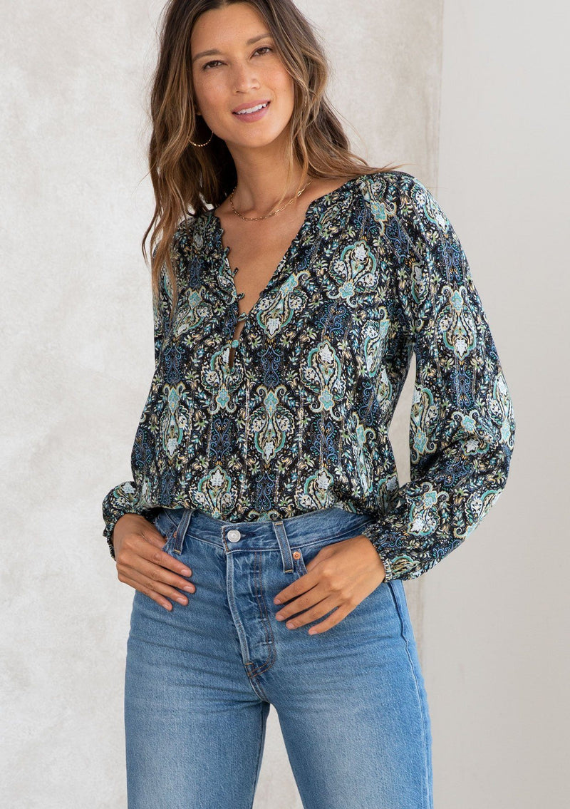 [Color: Black/Teal] A model wearing a black and teal paisley print peasant blouse with gold lurex thread details. With long voluminous sleeves, a self covered button front, and a button loop trimmed v neckline. 