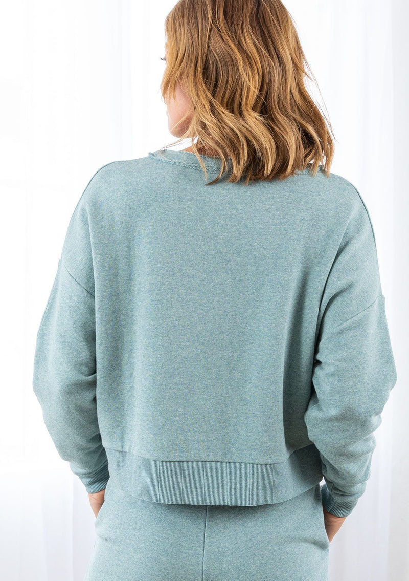 [Color: Sea Blue] A model wearing a cropped sea blue pullover sweatshirt with a buttoned front detail. With long raglan sleeves and a round neckline with raw unfinished hem.