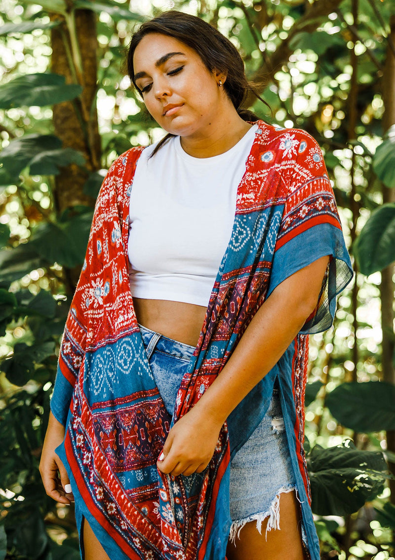 [Color: Blue/Brick] A model wearing a red and blue bohemian mid length kimono. Featuring half length kimono sleeves, side slits, an open front, and a multi colored pattern throughout.