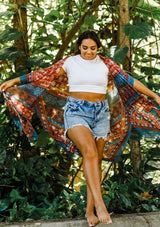 [Color: Blue/Brick] A model wearing a red and blue bohemian mid length kimono. Featuring half length kimono sleeves, side slits, an open front, and a multi colored pattern throughout.