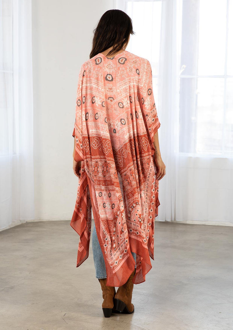 [Color: Brick/Coral] A model wearing a red and coral bohemian mid length kimono. Featuring half length kimono sleeves, side slits, an open front, and a multi colored pattern throughout.