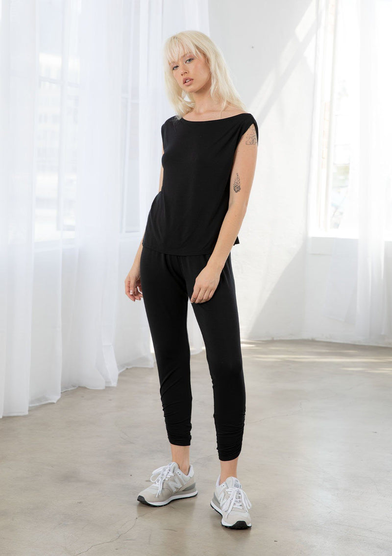 [Color: Black] A model wearing a classic black athleisure tank top. With a boat neckline, a gathered detail along the left shoulder, and a boxy fit.