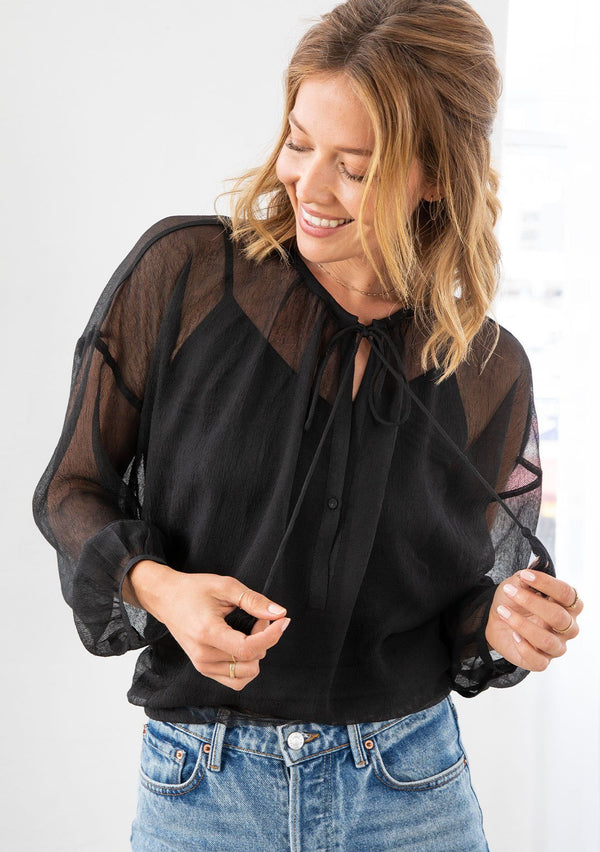 [Color: Black] A model wearing a sheer black peasant blouse. With long voluminous sleeves, a tassel tie neck, and front keyhole detail.