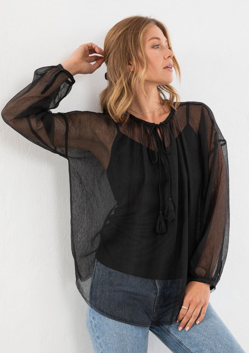 [Color: Black] A model wearing a sheer black peasant blouse. With long voluminous sleeves, a tassel tie neck, and front keyhole detail.