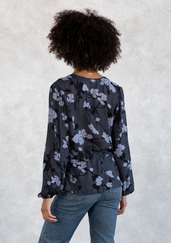 [Color: Slate/Grey] A model wearing a blue and grey floral print blouse. With a mini pom trimmed surplice neckline, voluminous long sleeves, an adjustable drawstring waist with side ties, and a peplum waist. 