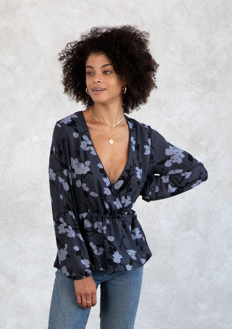 [Color: Slate/Grey] A model wearing a blue and grey floral print blouse. With a mini pom trimmed surplice neckline, voluminous long sleeves, an adjustable drawstring waist with side ties, and a peplum waist. 