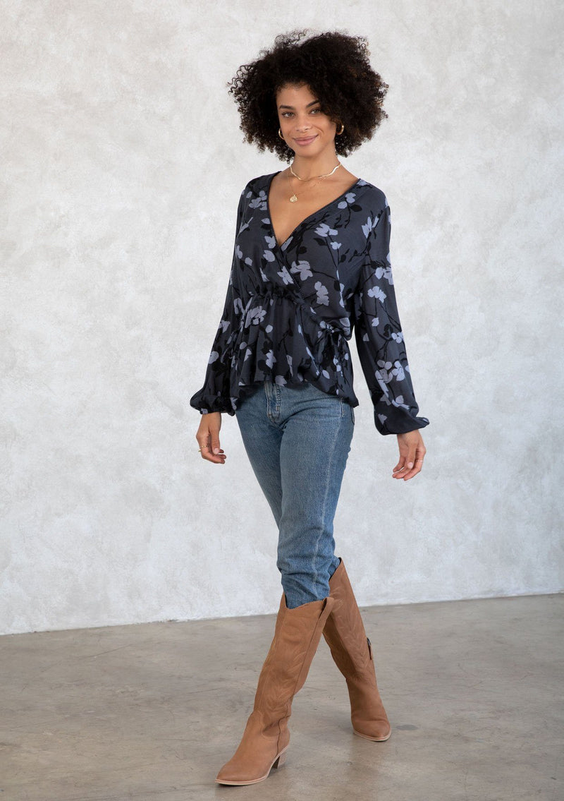 [Color: Slate/Grey] A model wearing a blue and grey floral print blouse. With a mini pom trimmed surplice neckline, voluminous long sleeves, an adjustable drawstring waist with side ties, and a peplum waist. 