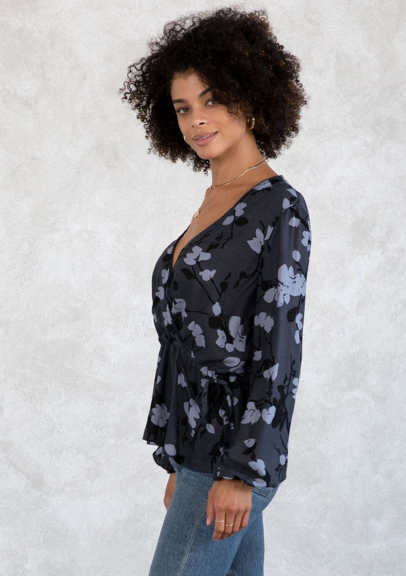 [Color: Slate/Grey] A model wearing a blue and grey floral print blouse. With a mini pom trimmed surplice neckline, voluminous long sleeves, an adjustable drawstring waist with side ties, and a peplum waist. 