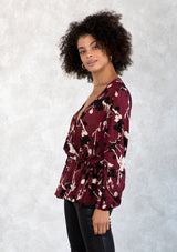 [Color: Merlot/Ivory] A model wearing a dark red and ivory floral print blouse. With a mini pom trimmed surplice neckline, voluminous long sleeves, an adjustable drawstring waist with side ties, and a peplum waist. 