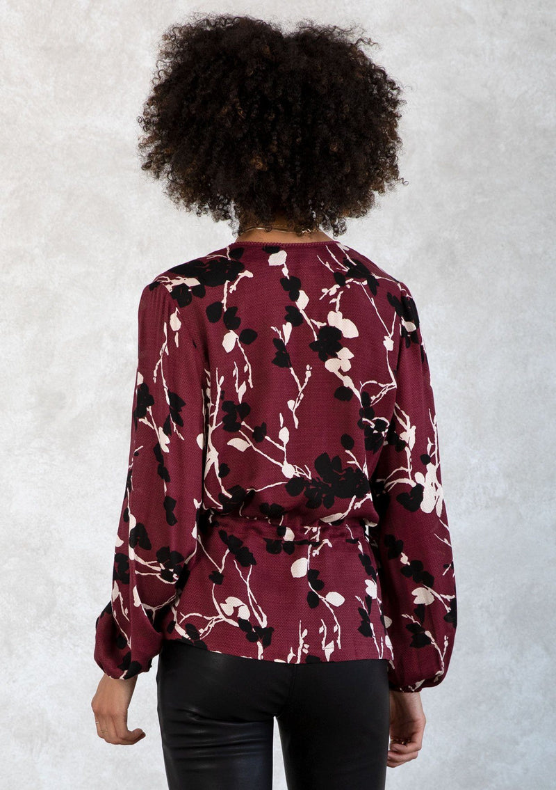 [Color: Merlot/Ivory] A model wearing a dark red and ivory floral print blouse. With a mini pom trimmed surplice neckline, voluminous long sleeves, an adjustable drawstring waist with side ties, and a peplum waist. 