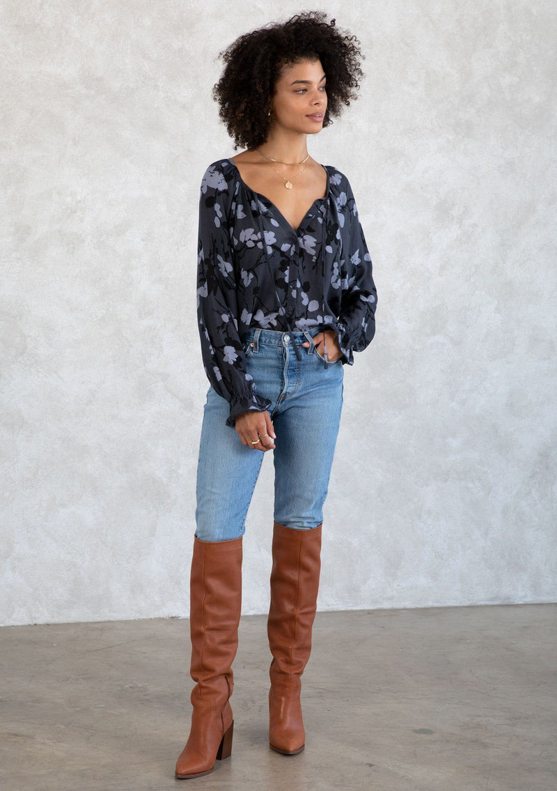 [Color: Slate/Grey] A model wearing a blue fall floral print bohemian blouse. With a self covered button front, a split neckline with ties, voluminous long sleeves, and a flounce elastic wrist cuffs with a mini pom trim.