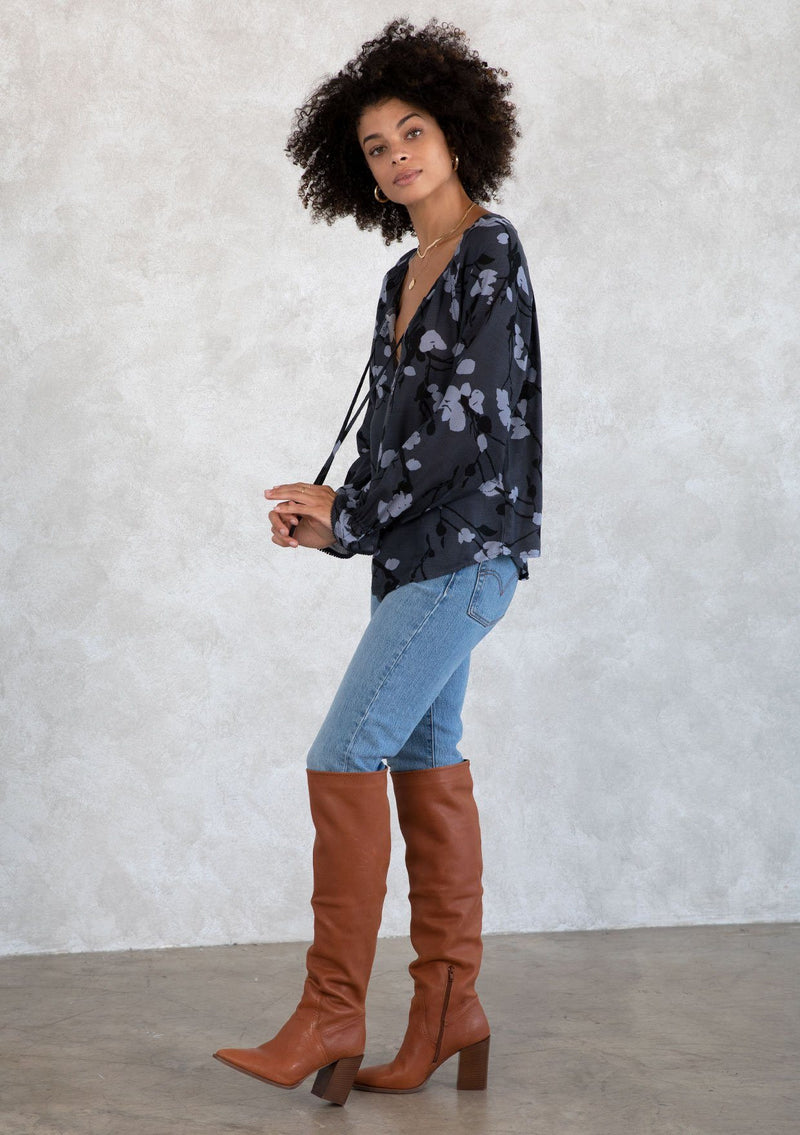 [Color: Slate/Grey] A model wearing a blue fall floral print bohemian blouse. With a self covered button front, a split neckline with ties, voluminous long sleeves, and a flounce elastic wrist cuffs with a mini pom trim.