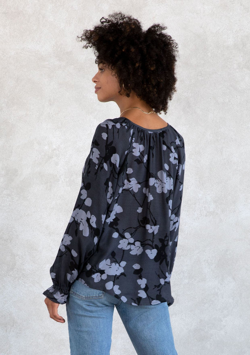 [Color: Slate/Grey] A model wearing a blue fall floral print bohemian blouse. With a self covered button front, a split neckline with ties, voluminous long sleeves, and a flounce elastic wrist cuffs with a mini pom trim.