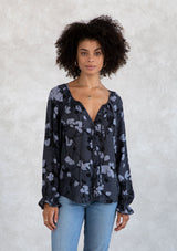 [Color: Slate/Grey] A model wearing a blue fall floral print bohemian blouse. With a self covered button front, a split neckline with ties, voluminous long sleeves, and a flounce elastic wrist cuffs with a mini pom trim.