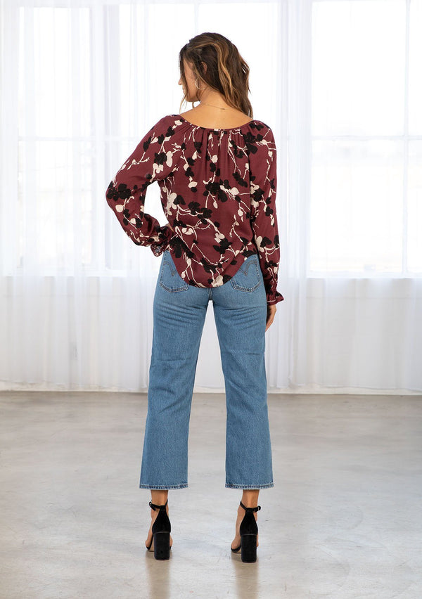 [Color: Merlot/Ivory] A model wearing a burgundy red and ivory fall floral print bohemian blouse. With a self covered button front, a split neckline with ties, voluminous long sleeves, and a flounce elastic wrist cuffs with a mini pom trim. 