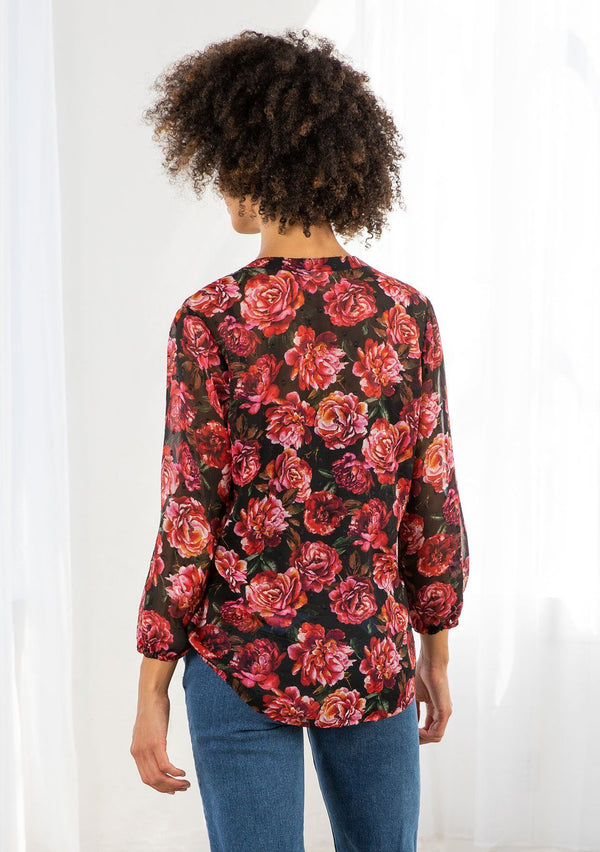 [Color: Black/Magenta] A model wearing a sheer red and black floral print blouse. With long voluminous sleeves, elastic wrist cuffs, a self covered button front, a split neckline with ties, lace trim detail, and clip dot details throughout. 