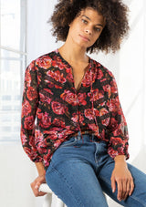 [Color: Black/Magenta] A model wearing a sheer red and black floral print blouse. With long voluminous sleeves, elastic wrist cuffs, a self covered button front, a split neckline with ties, lace trim detail, and clip dot details throughout. 