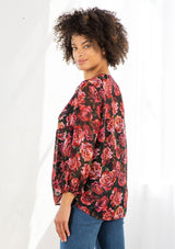 [Color: Black/Magenta] A model wearing a sheer red and black floral print blouse. With long voluminous sleeves, elastic wrist cuffs, a self covered button front, a split neckline with ties, lace trim detail, and clip dot details throughout. 