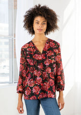 [Color: Black/Magenta] A model wearing a sheer red and black floral print blouse. With long voluminous sleeves, elastic wrist cuffs, a self covered button front, a split neckline with ties, lace trim detail, and clip dot details throughout. 