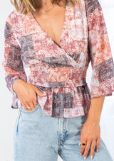 [Color: Natural/Rose] A model wearing a bohemian blouse in a red paisley patchwork print. With three quarter length bell sleeves, a faux surplice neckline, and a smocked elastic waist. 