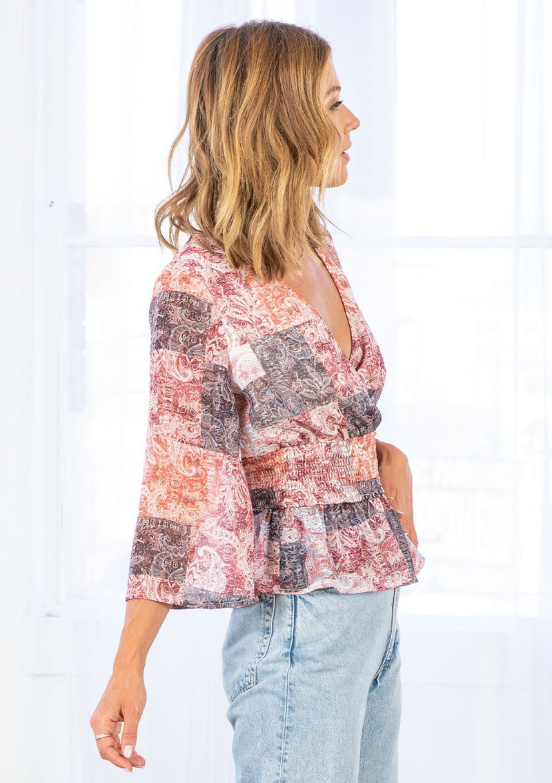 [Color: Natural/Rose] A model wearing a bohemian blouse in a red paisley patchwork print. With three quarter length bell sleeves, a faux surplice neckline, and a smocked elastic waist. 