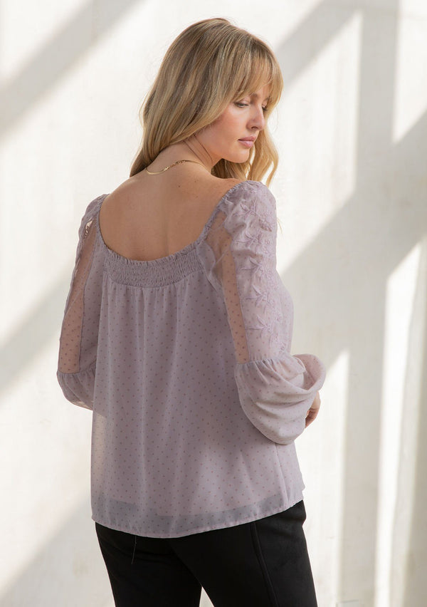 [Color: Vintage Lilac] A model wearing a romantic square neck blouse in a lilac purple dot chiffon. With a long voluminous lantern sleeve with embroidered dot mesh detail and a billowy relaxed fit.